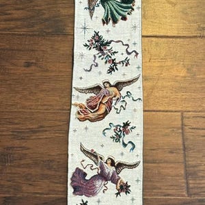 May include: A long, vertical woven wall hanging featuring a cream-colored background with five distinct angel figures in various poses and colorful robes. The design includes small floral accents, ribbons, and star motifs scattered between the angels.