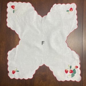 May include: White linen tea towel with a scalloped edge and red embroidery of strawberries. The towel has the letter "F" embroidered in the center.