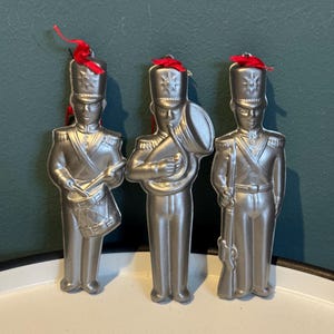 Op de afbeelding: Drie zilverkleurige muziekgroepornamenten. Elk ornament toont een uniform figuur met een rood lint aan de bovenkant. Eén figuur houdt een trommel vast, een ander een tuba en de derde een geweer. De ornamenten staan op een wit dienblad.