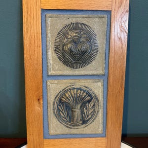 May include: A framed artwork featuring two square ceramic tiles with blue and gray designs. The top tile has a heart-shaped design, and the bottom tile has a wheat sheaf design. The frame is made of light brown wood, and the background is a light blue.