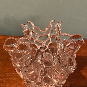 Puede incluir: Un cuenco de vidrio transparente con un diseño texturizado único. El exterior del cuenco presenta un patrón orgánico y burbujeante, y el borde tiene una forma ondulada y con volantes. El vidrio es transparente, permitiendo el paso de la luz, y se asienta sobre una base circular.