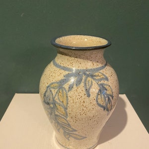 May include: A ceramic vase with a speckled cream-colored glaze and a blue hand-painted leaf pattern. The vase features a rounded body, a narrow neck, and a dark blue rim. The surface has a glossy finish with visible brown speckles throughout.