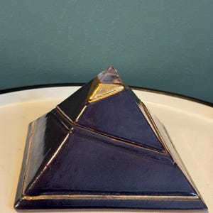Può includere: Oggetto decorativo a forma di piramide blu scuro con finiture dorate. La parte superiore della piramide presenta una punta di cristallo trasparente. Appoggiato su una superficie bianca.