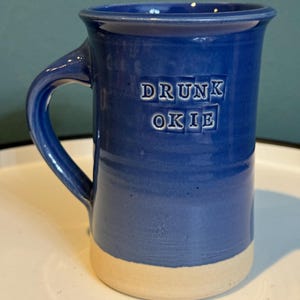 May include: A blue ceramic mug with a white rim and handle. The mug has the words "DRUNK OKIE" printed on the front in white.