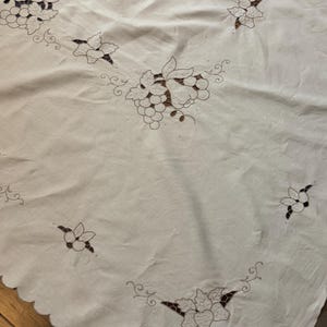 Puede incluir: Un mantel blanco con intrincados bordados calados y detalles en hilo marrón. El diseño muestra racimos de uvas, peras y hojas dispuestos en un patrón repetitivo sobre la tela, que presenta un acabado de borde festoneado.