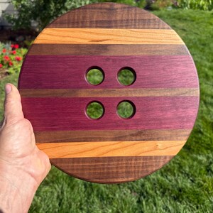 May include: A round wooden button-shaped object with four holes. The button is made of alternating stripes of dark brown, orange, and purple wood. The object is held by a hand against a background of green grass and foliage.