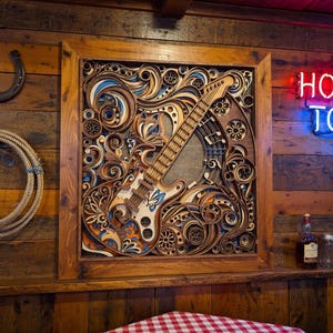 May include: A wooden framed, layered art piece featuring a guitar design with intricate details in shades of brown, blue, and orange. The neon sign above reads "HONKY TONK". A horseshoe and rope are on the wooden wall.