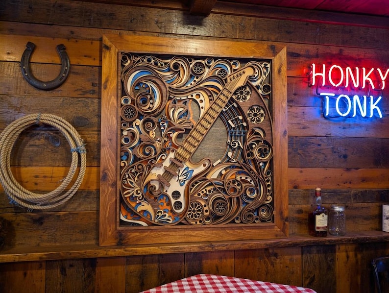 May include: A wooden framed, layered art piece featuring a guitar design with intricate details in shades of brown, blue, and orange. The neon sign above reads "HONKY TONK". A horseshoe and rope are on the wooden wall.