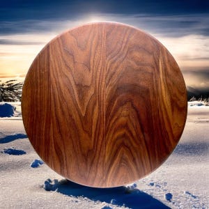 May include: A large, round wooden cutting board with a rich, brown wood grain pattern. The board is set against a snowy, winter landscape with a bright sky in the background. The board appears to be approximately 18 inches in diameter.