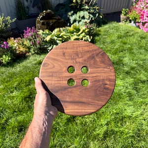 May include: A large, circular wooden button with four holes, held by a person's hand. The button is made of dark brown wood and has a visible wood grain pattern. The background is a lush green lawn with flowers.