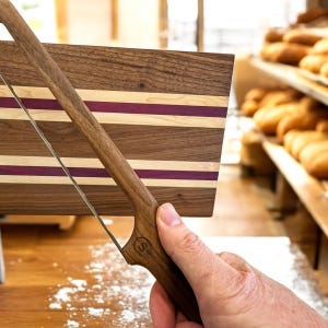 Puede incluir: Una tabla de cortar de madera con rayas moradas y de madera clara y un cuchillo para pan con mango de madera. El cuchillo tiene una hoja dentada y el mango tiene la letra "S" grabada.