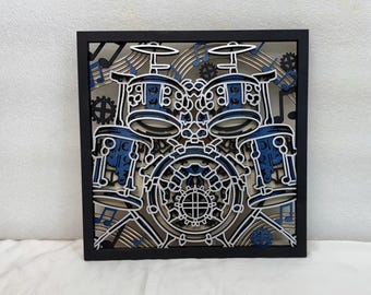 May include: A layered wooden art piece featuring a drum set design. The artwork has a black frame, with blue and white accents. Musical notes and gears are incorporated into the design, creating a detailed and artistic representation of a drum kit.