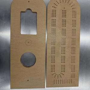 May include: Two unfinished wooden cribbage board pieces. One piece has a large circular hole and a rectangular cutout. The other piece has numerous small holes for peg placement. Both pieces are light brown and have an arched top.