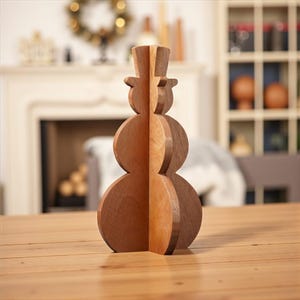 May include: A wooden snowman decoration with a top hat, constructed from layered wood pieces. The snowman is brown and tan, with a simple, geometric design. It is standing on a wooden table.