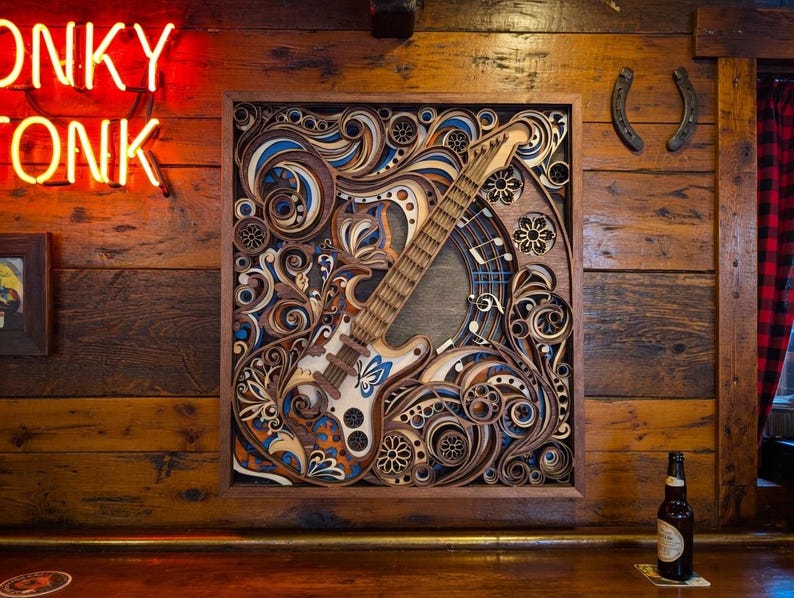 May include: A wooden, layered art piece featuring a guitar design with intricate details in shades of brown, blue, and white. The artwork is mounted on a wooden wall, with a neon "ONKY TONK" sign visible. A beer bottle sits on a wooden bar.