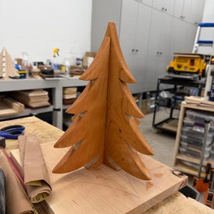 May include: A handcrafted wooden Christmas tree with a natural finish. The tree is made of two interlocking pieces and sits on a square wooden base. The tree has multiple layers of branches and a pointed top. The wood has a warm, reddish-brown hue.
