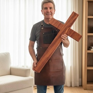 May include: A large, handcrafted wooden cross with a rich, reddish-brown stain. The cross is held by a person wearing a brown leather apron. The cross appears to be made of solid wood and has a simple, yet elegant design.
