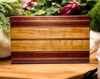 Może przedstawiać: Prostokątna drewniana deska do krojenia z poziomymi pasami o różnej szerokości i kolorach. Deska posiada naprzemienne pasma usłojenia drewna w odcieniach głębokiego czerwonawego brązu, jasnego złocistego brązu oraz ciemnego brązu.