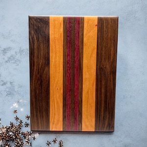 May include: A rectangular wooden cutting board with a striped design. The board features alternating vertical stripes of dark brown, orange, and red-purple wood. The cutting board is made of hardwood and has a smooth, polished finish.
