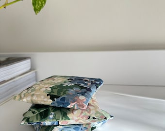 May include: A stack of three square pillows with a floral pattern in shades of green, blue, and pink. The pillows are stacked on a white surface, with a trailing green plant in the background.