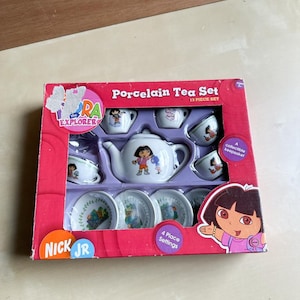 May include: A 13-piece porcelain tea set in a pink box featuring Dora the Explorer. The set includes a teapot, four cups, saucers, and the text "Porcelain Tea Set" and "4 Place Settings".