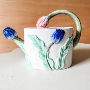 May include: White ceramic watering can with a green handle shaped like a tulip stem. The can is decorated with three tulips, one pink and two blue.
