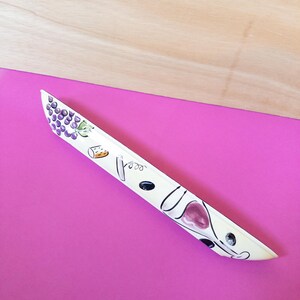 Könnte beinhalten: Weiße Keramik-Servierplatte mit einer Schwarzweiß-Illustration von Trauben, Weingläsern und Korkenziehern. Die Platte liegt auf einer rosa Oberfläche.