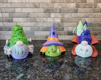 May include: Three Halloween gnomes, one wearing a green hat, one wearing a purple hat with a star, and one wearing a purple hat with a green and white striped shirt.