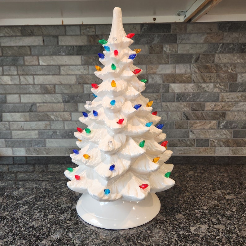 K&ouml;nnte beinhalten: Ein wei&szlig;er Keramik-Weihnachtsbaum mit bunten Lichtern. Der Baum ist mit roten, gr&uuml;nen, blauen und gelben Lichtern geschm&uuml;ckt. Der Baum steht auf einer schwarzen Arbeitsplatte.