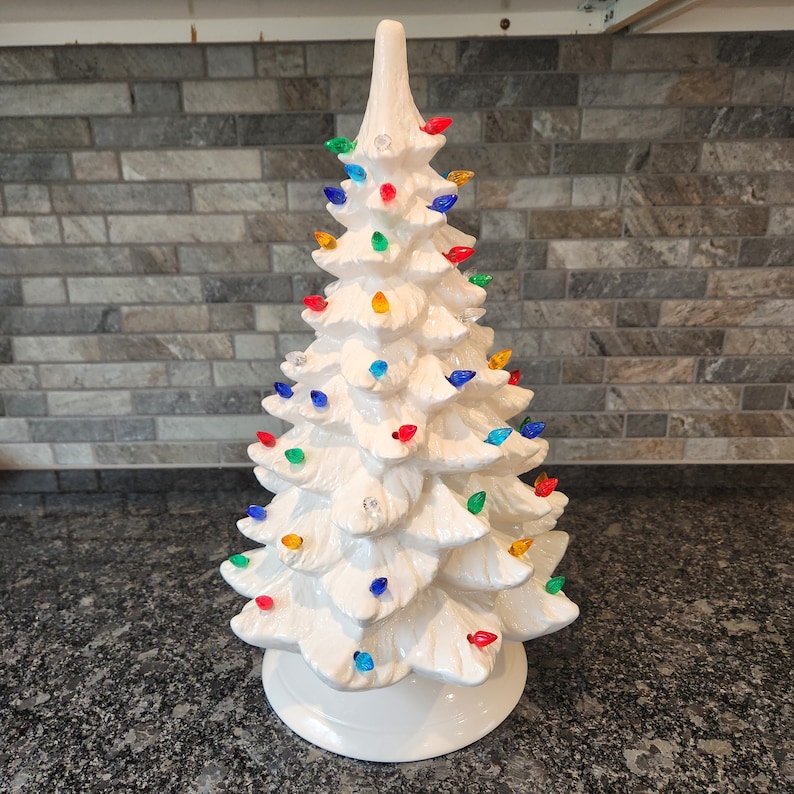 K&ouml;nnte beinhalten: Ein wei&szlig;er Keramik-Weihnachtsbaum mit bunten Lichtern. Der Baum ist mit roten, gr&uuml;nen, blauen und gelben Lichtern geschm&uuml;ckt. Der Baum steht auf einer schwarzen Arbeitsplatte.