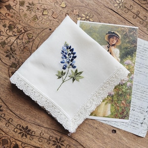 May include: A square white handkerchief featuring an embroidered blue lupine flower with green leaves in the center and a delicate white lace trim along the edges. The fabric is smooth and the floral embroidery displays shades of light and dark blue.