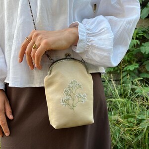 May include: A cream-coloured crossbody bag with floral embroidery. The bag features a metal clasp and chain strap. The bag is held by a person wearing a white blouse and brown skirt.
