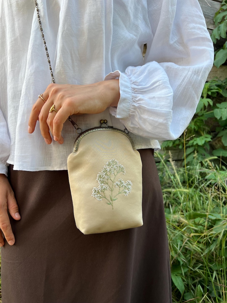 May include: A cream-coloured crossbody bag with floral embroidery. The bag features a metal clasp and chain strap. The bag is held by a person wearing a white blouse and brown skirt.