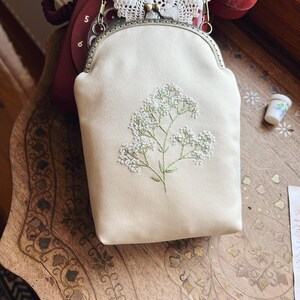 May include: A cream-coloured handbag featuring floral embroidery. The bag has a metal clasp and a chain strap. The embroidered flowers are white with green stems. The bag rests on a wooden surface.