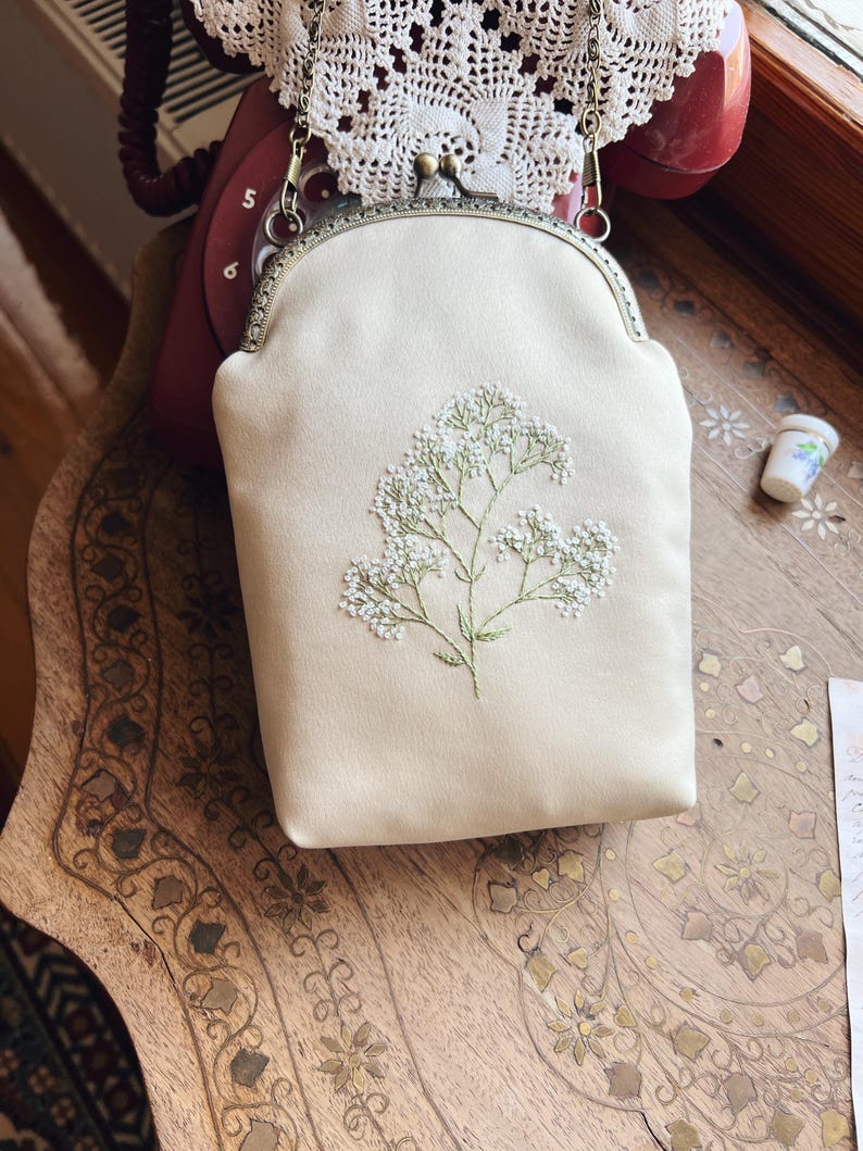 May include: A cream-coloured handbag featuring floral embroidery. The bag has a metal clasp and a chain strap. The embroidered flowers are white with green stems. The bag rests on a wooden surface.