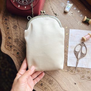 May include: A cream satin clutch bag with a metal clasp and chain strap. The rectangular purse has a rounded top. The background features a vintage red rotary phone, sewing supplies, and a decorative wooden table.