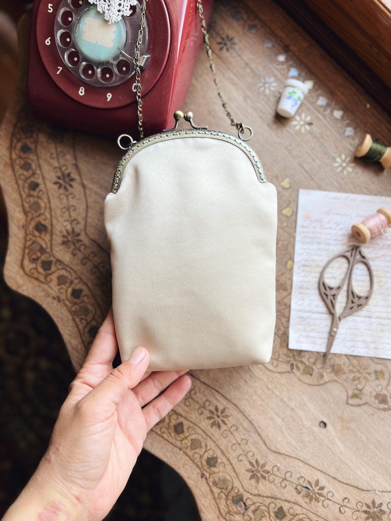 May include: A cream satin clutch bag with a metal clasp and chain strap. The rectangular purse has a rounded top. The background features a vintage red rotary phone, sewing supplies, and a decorative wooden table.