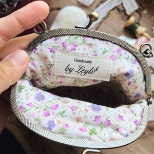 May include: An open, vintage-style coin purse with a floral interior. The purse has a metal frame and clasp. The interior fabric features a pattern of pink, purple, and green flowers on a cream background. The label reads "Handmade by Leyli".