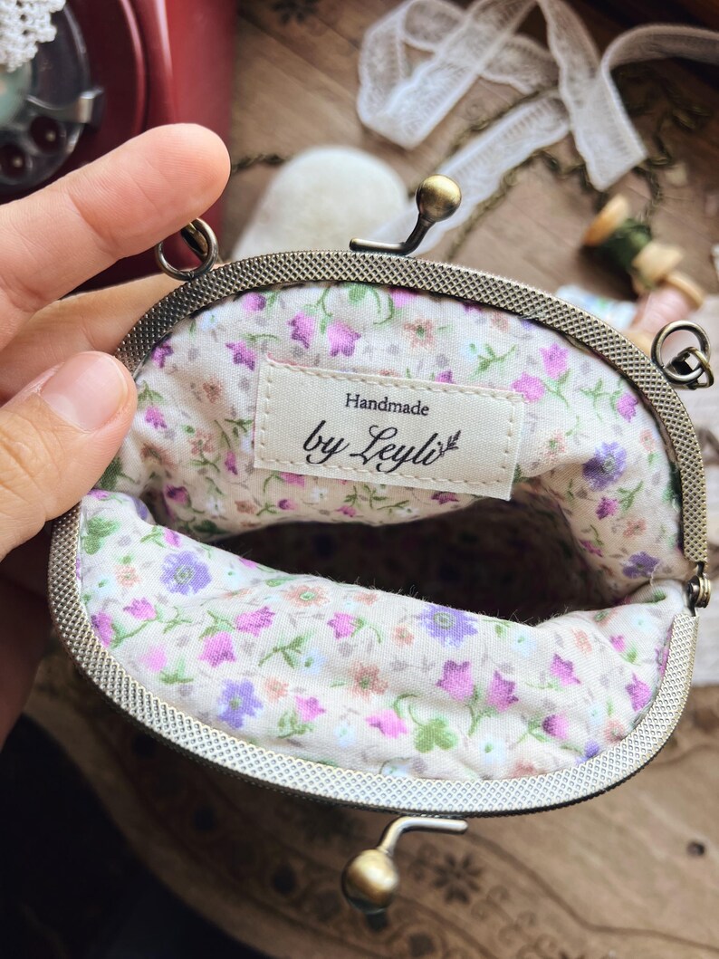 May include: An open, vintage-style coin purse with a floral interior. The purse has a metal frame and clasp. The interior fabric features a pattern of pink, purple, and green flowers on a cream background. The label reads "Handmade by Leyli".