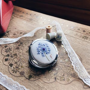 Puede incluir: Un espejo compacto plateado con un diseño de bordado floral azul. El espejo está abierto, revelando el detalle bordado. Una cinta de encaje blanco, un carrete de hilo y un dedal también son visibles sobre una superficie de madera.