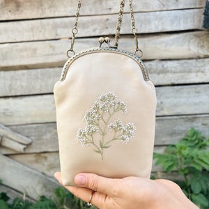 May include: A beige clutch bag with a floral embroidery design. The bag has a metal clasp and a chain strap. The embroidered design features white flowers and green stems. The bag is held up by a hand.