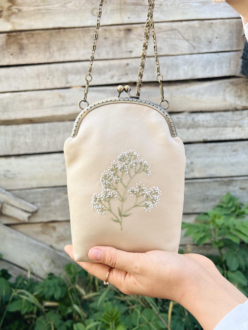 May include: A beige clutch bag with a floral embroidery design. The bag has a metal clasp and a chain strap. The embroidered design features white flowers and green stems. The bag is held up by a hand.