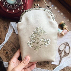 May include: A cream-coloured satin clutch bag with a floral embroidery design. The bag has a metal clasp and a chain strap. The embroidery features small white flowers and green stems. The bag is held by a hand.
