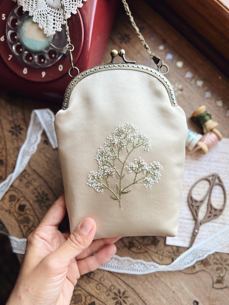 May include: A cream-coloured satin clutch bag with a floral embroidery design. The bag has a metal clasp and a chain strap. The embroidery features small white flowers and green stems. The bag is held by a hand.