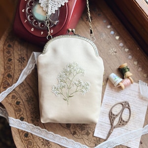 May include: A cream-coloured, vintage-style purse with a floral embroidery design. The purse has a metal clasp and a chain strap. The image also shows a red rotary phone, lace trim, and sewing notions on a wooden surface.