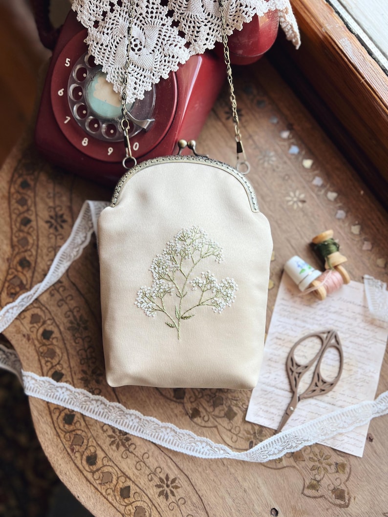 May include: A cream-coloured, vintage-style purse with a floral embroidery design. The purse has a metal clasp and a chain strap. The image also shows a red rotary phone, lace trim, and sewing notions on a wooden surface.