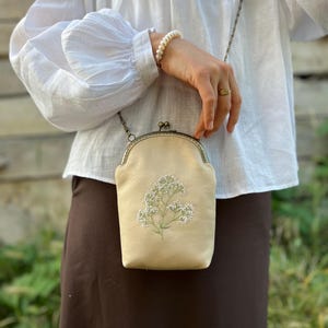 May include: A beige crossbody bag with floral embroidery. The bag features a metal clasp and chain strap. The bag is approximately 18 cm tall and 13 cm wide. The bag is held by a person wearing a white blouse and a brown skirt.