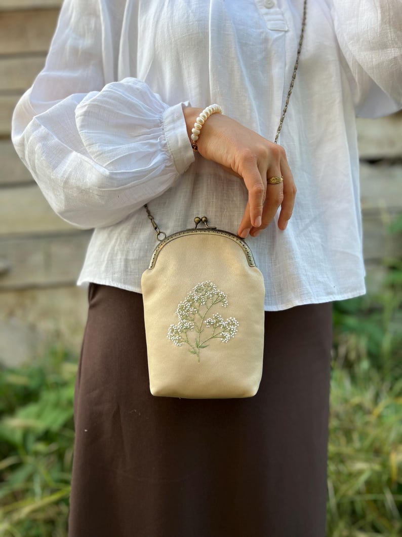 May include: A beige crossbody bag with floral embroidery. The bag features a metal clasp and chain strap. The bag is approximately 18 cm tall and 13 cm wide. The bag is held by a person wearing a white blouse and a brown skirt.
