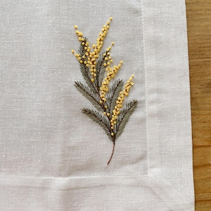 Peut inclure: Un set de table en lin blanc avec une broderie détaillée d'une branche de mimosa. Le mimosa présente des grappes de fleurs jaunes et des feuilles vertes et marron. Le set de table a une bordure simple.