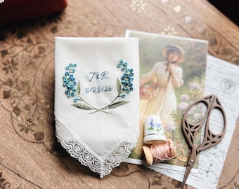 May include: A white handkerchief with embroidered blue flowers and the initials "T&K" with the date "8/28/26". The handkerchief has a delicate lace trim. Other items include a vintage postcard, scissors, and a thimble.